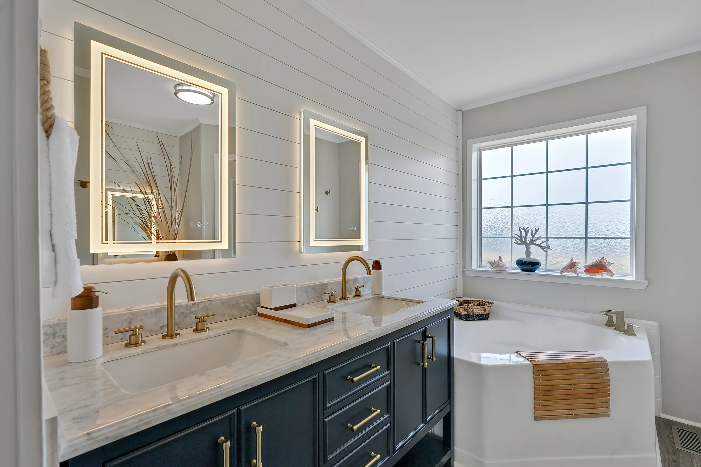 Bathroom with two illuminated mirrors above a double vanity beside a bathtub and window