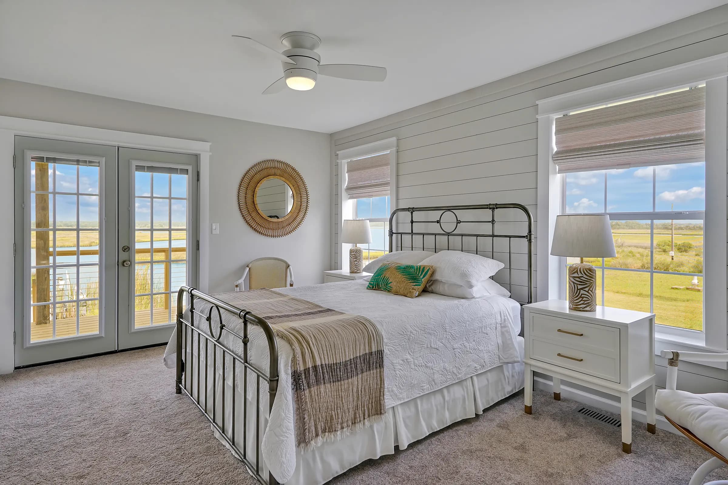 Primary bedroom with a metal-framed bed in white bedding, French doors, a round rattan mirror and windows overlooking marsh grass and water