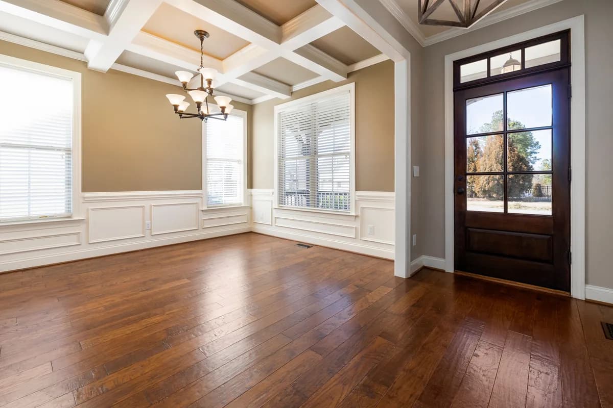 Empty living room with hardwood floors, large windows, white wall panels and a coffered ceiling